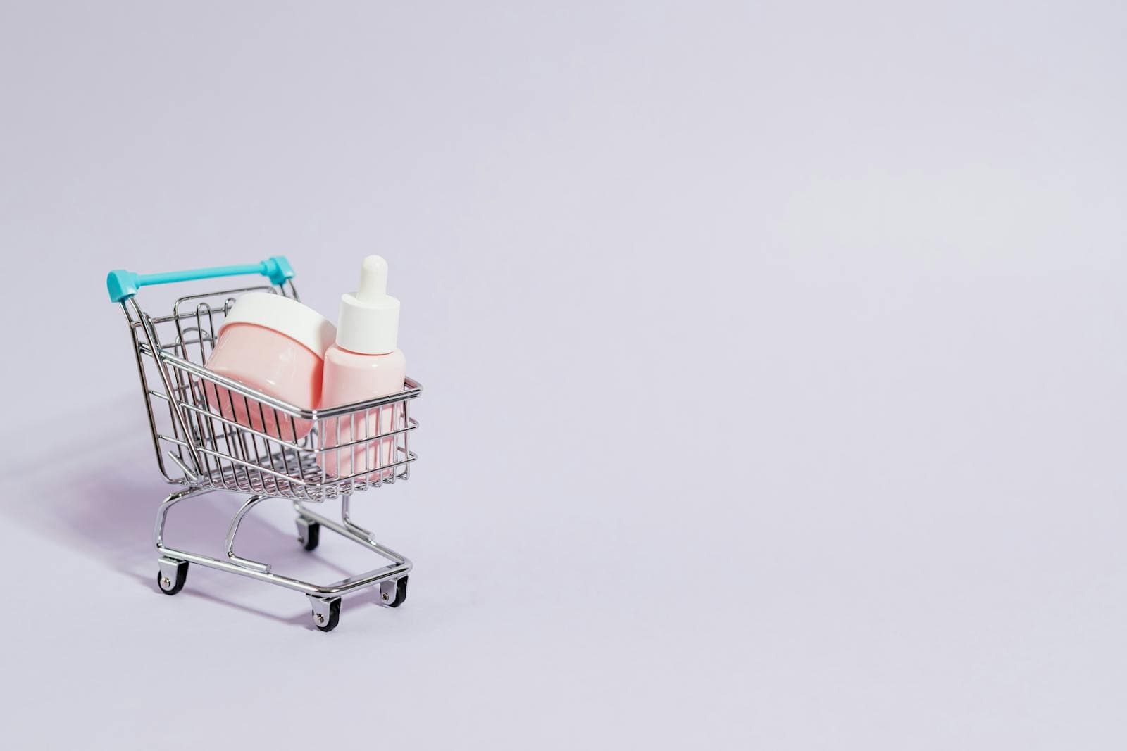Small shopping cart holding pink skincare products on a pastel background.