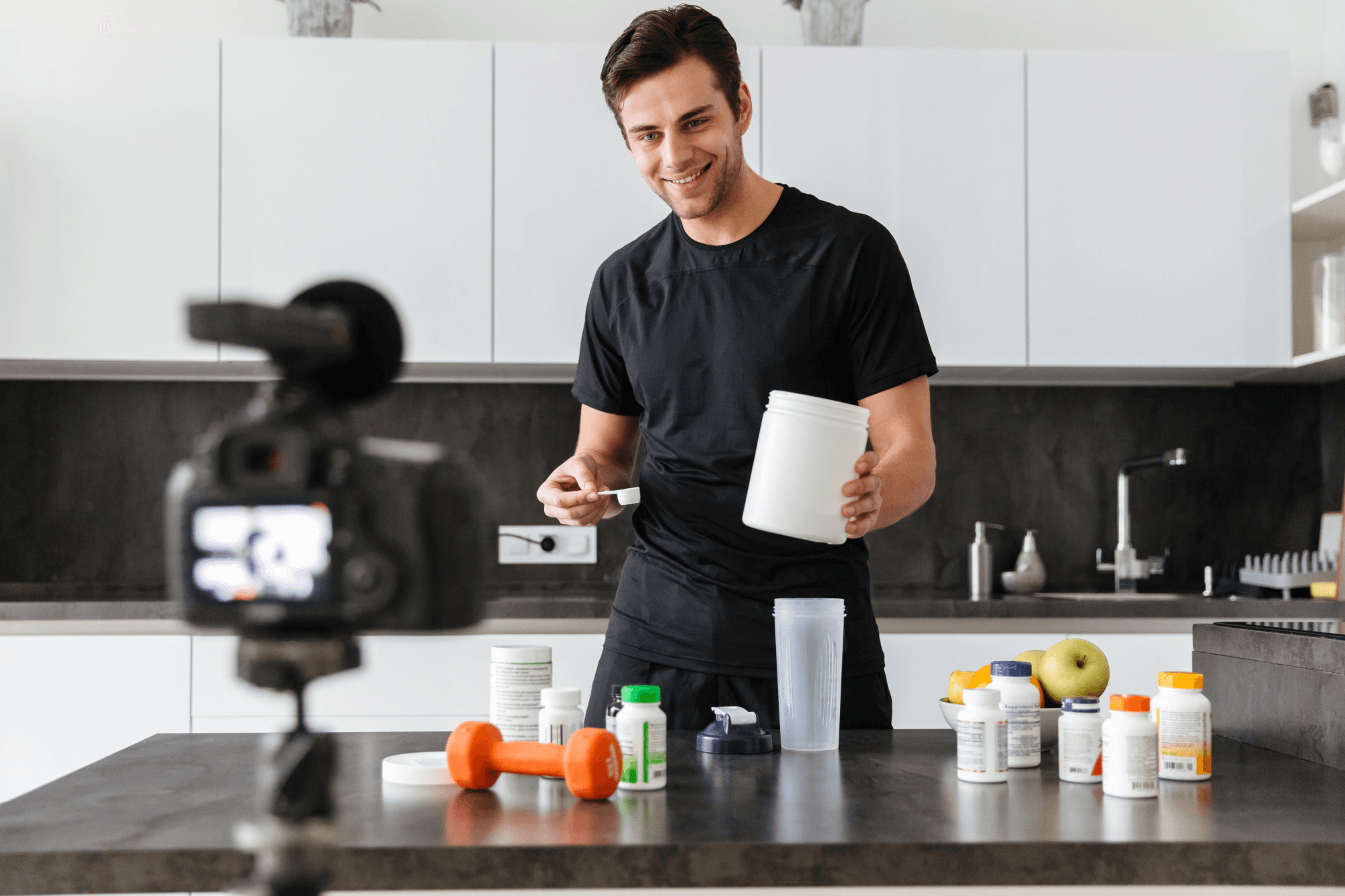 A man filming a video while holding a tub of supplement powder surrounded by bottles on a kitchen counter