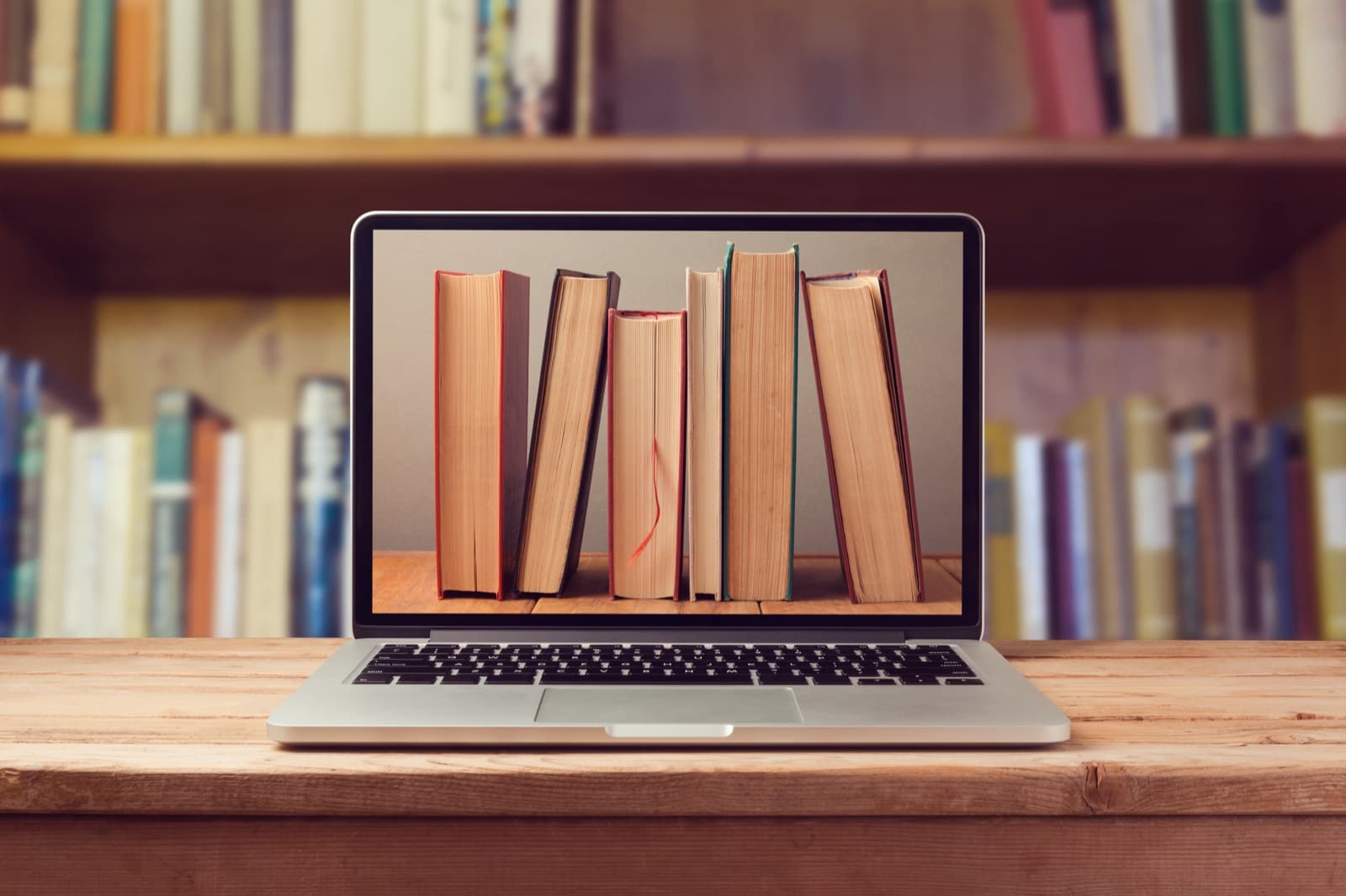 Laptop on a wooden desk displaying a row of books, with a full bookshelf in the background
