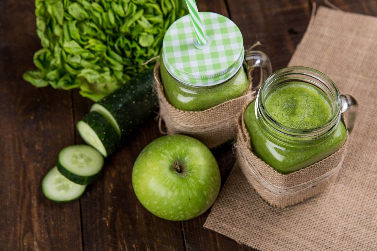 Green smoothie in mason jars with a green apple, cucumber and leafy greens on a wooden table