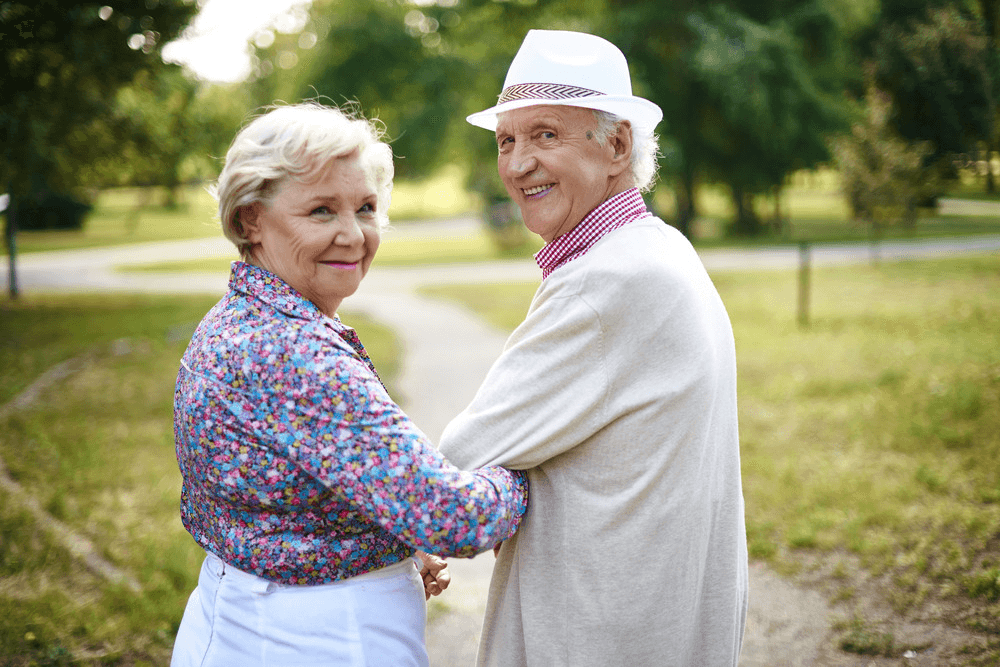 An elderly couple smiling and walking arm in arm through a park