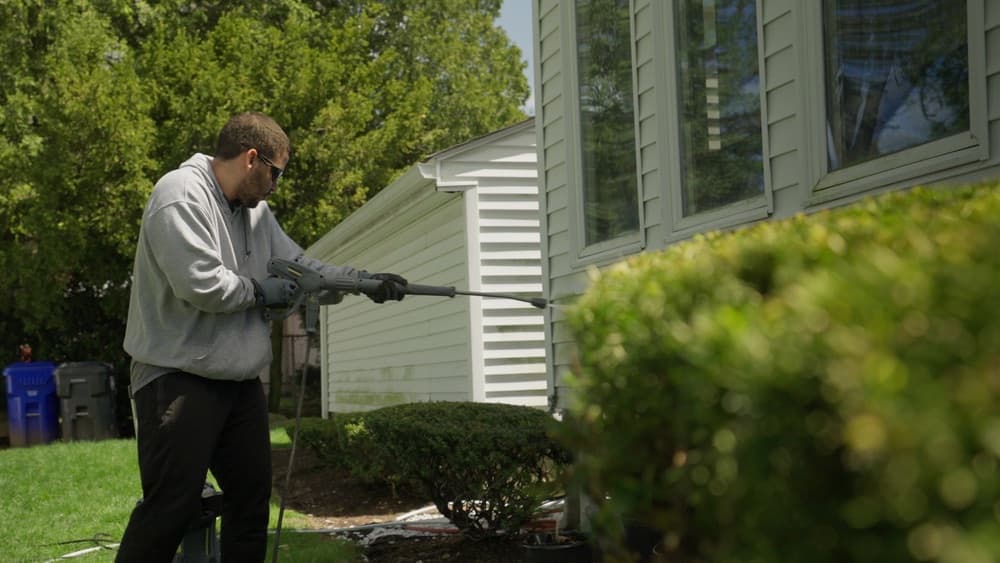 Homeowner pressure washing house siding beside a green lawn