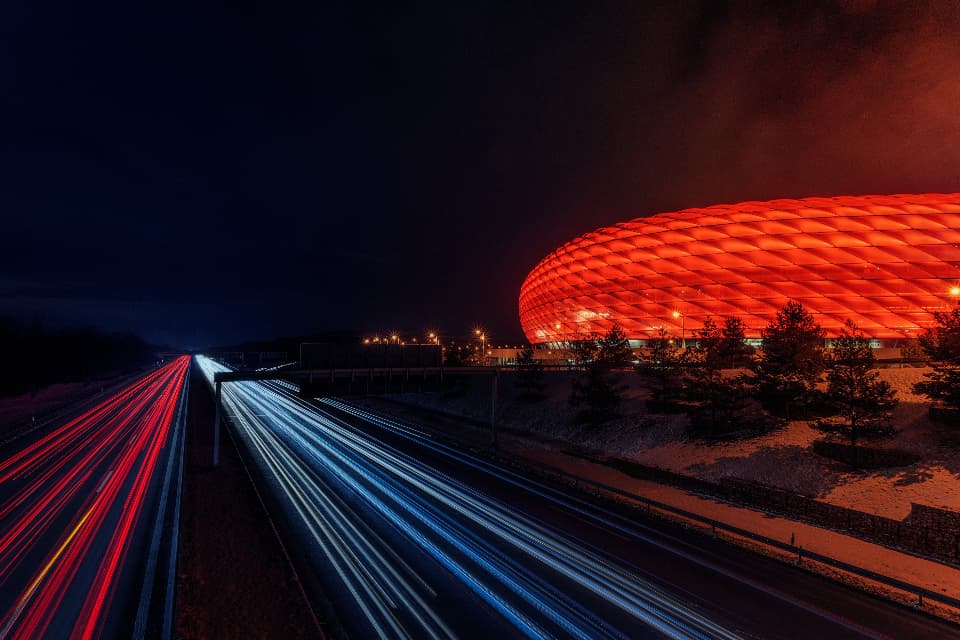 Illuminated sports stadium at night beside a motorway with light trails