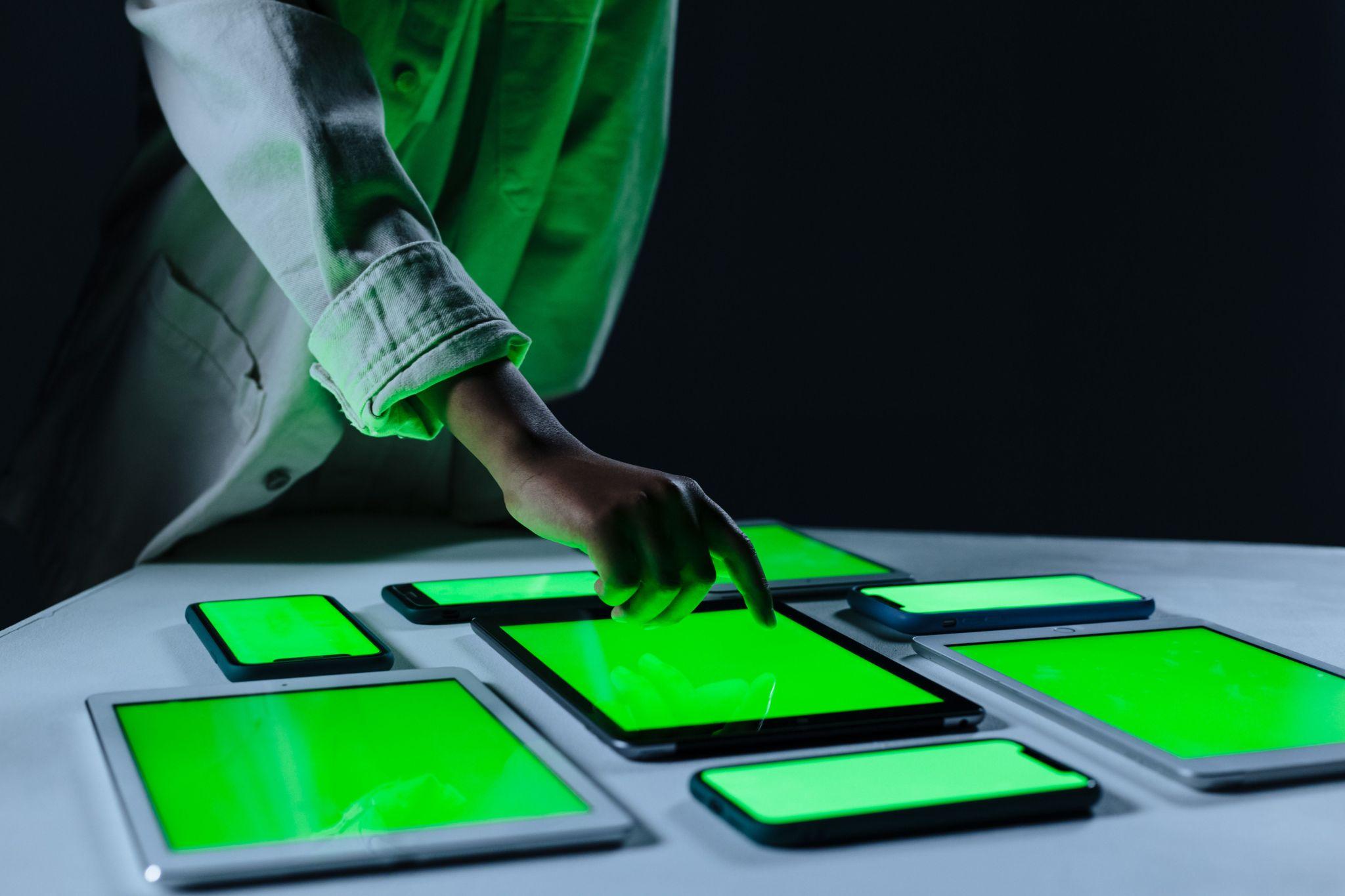 A hand touching one of several glowing tablet and phone screens on a table
